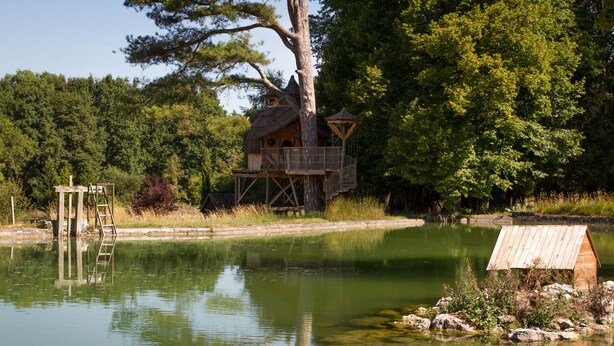 Ein hölzernes Baumhaus mit Plattform und Leiter, an einem ruhigen Teich gelegen, umgeben von Bäumen.