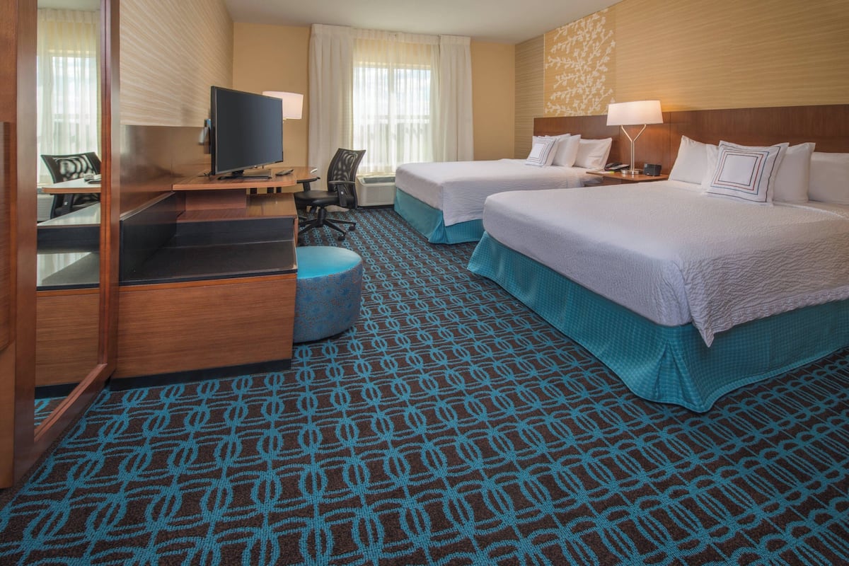 Ein Hotelzimmer mit zwei Betten, einem Schreibtisch mit Fernseher, einem Sessel und einem gemusterten Teppich.