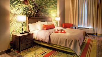 Una habitación de hotel con una cama grande, dos mesitas de noche con lámparas, una silla, un ventanal con cortinas y un mural en la pared.