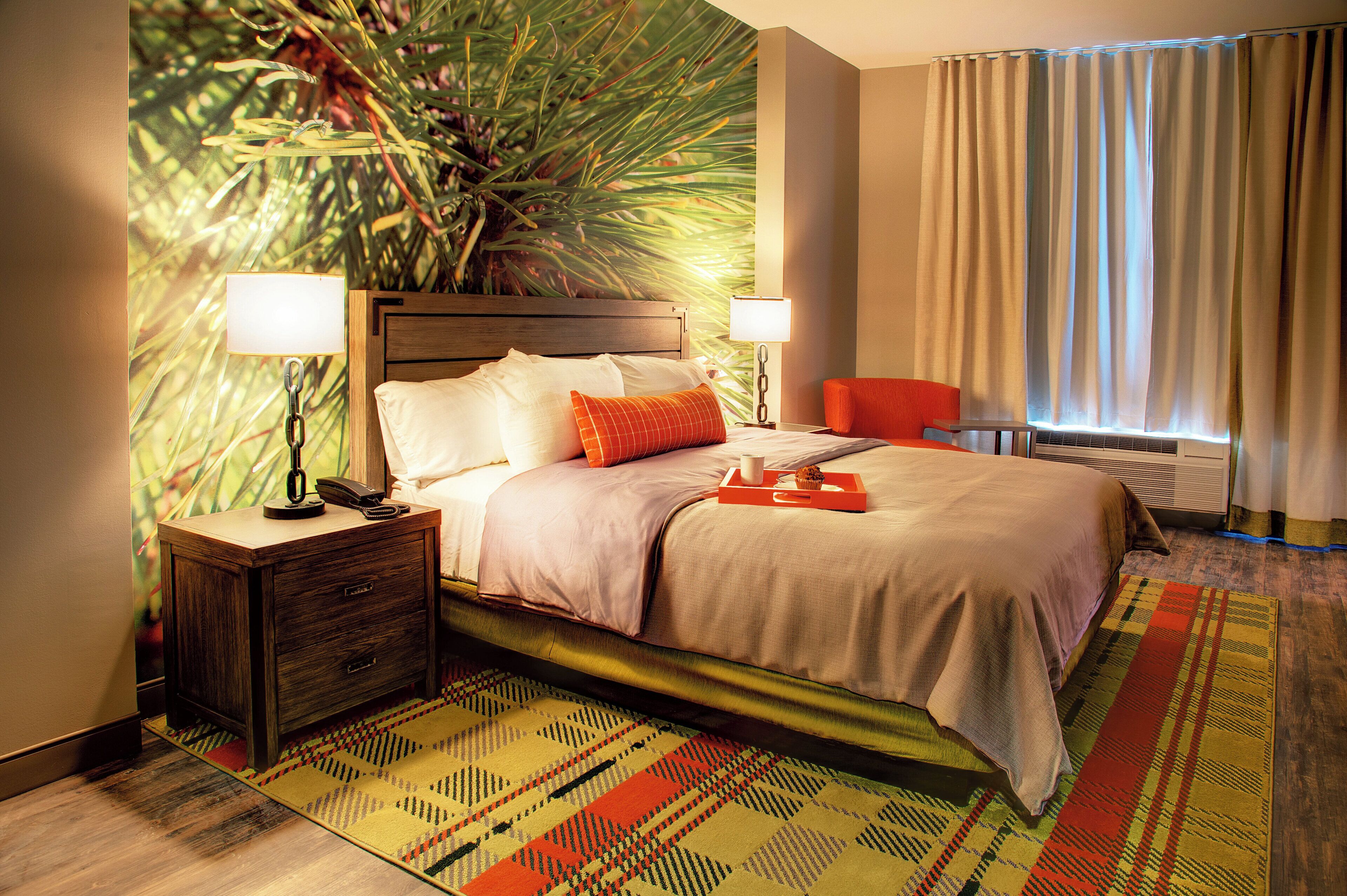 Une chambre d’hôtel comprenant un lit, des tables de chevet, une chaise, une fenêtre avec des rideaux et un mur orné d’une fresque représentant des plantes.
