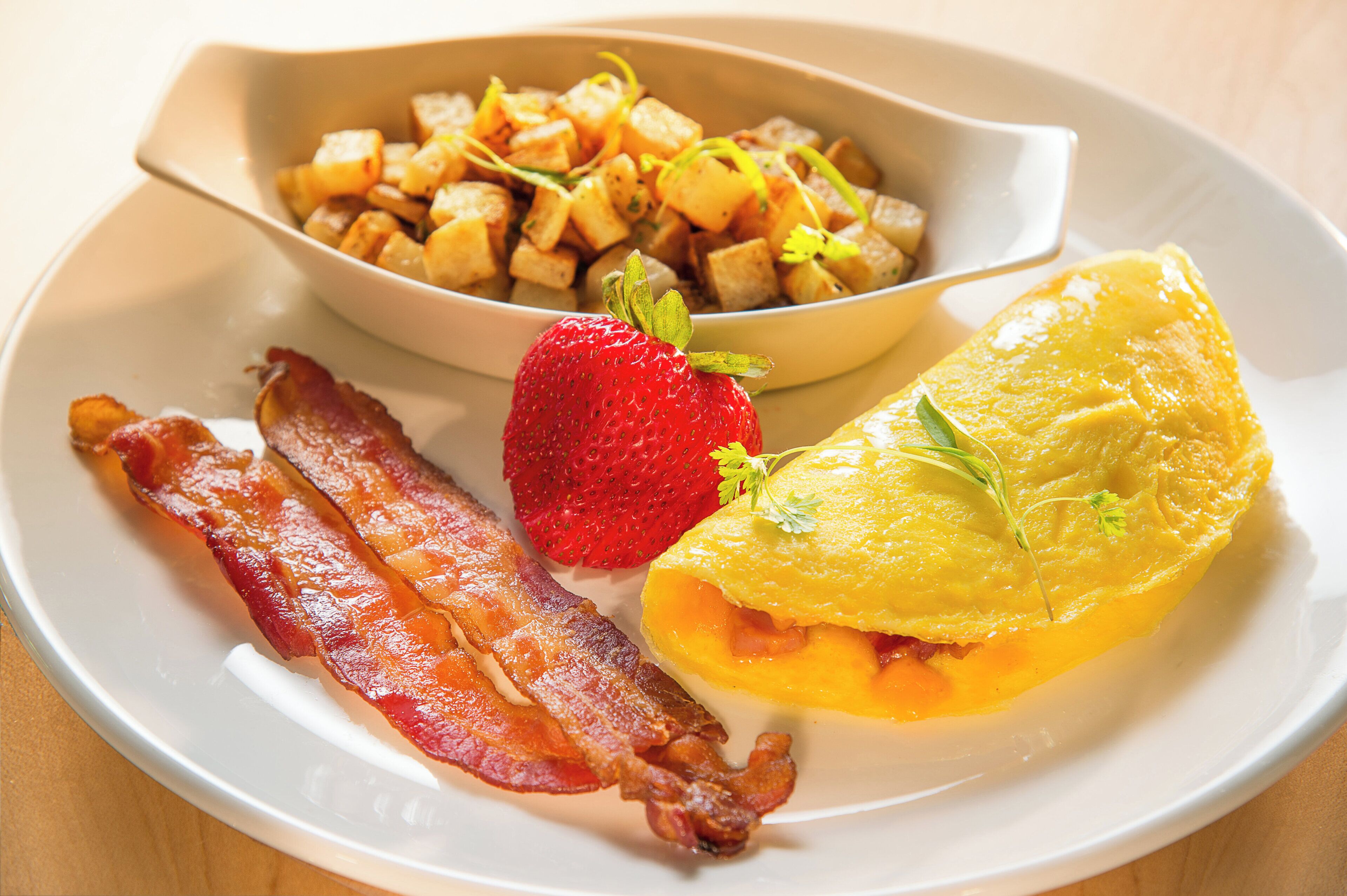 A plate with bacon, a strawberry, an omelette, and diced potatoes.