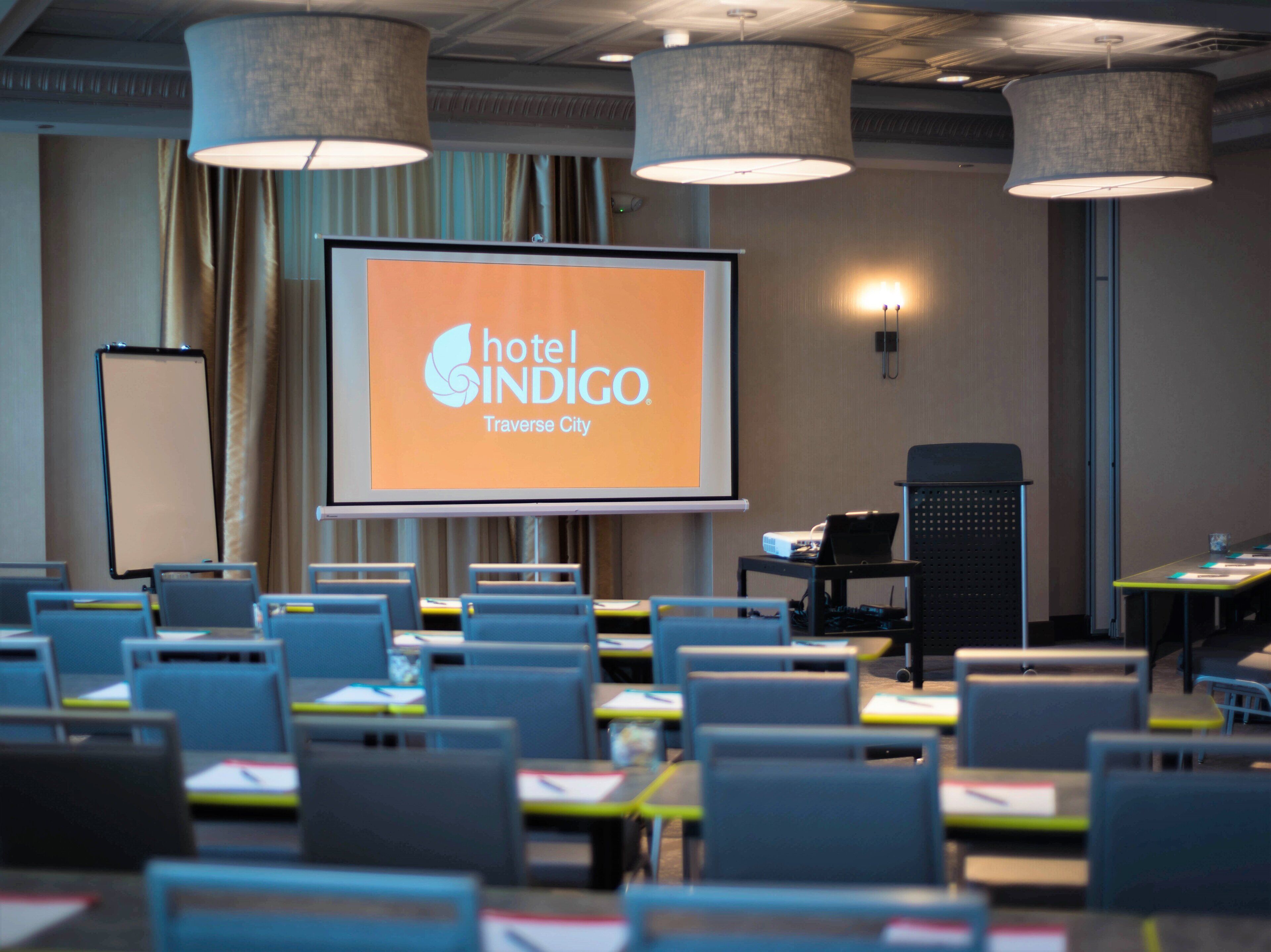 A conference room with a projection screen displaying 'hotel INDIGO Traverse City' branding, multiple tables with chairs, and a speaker system.