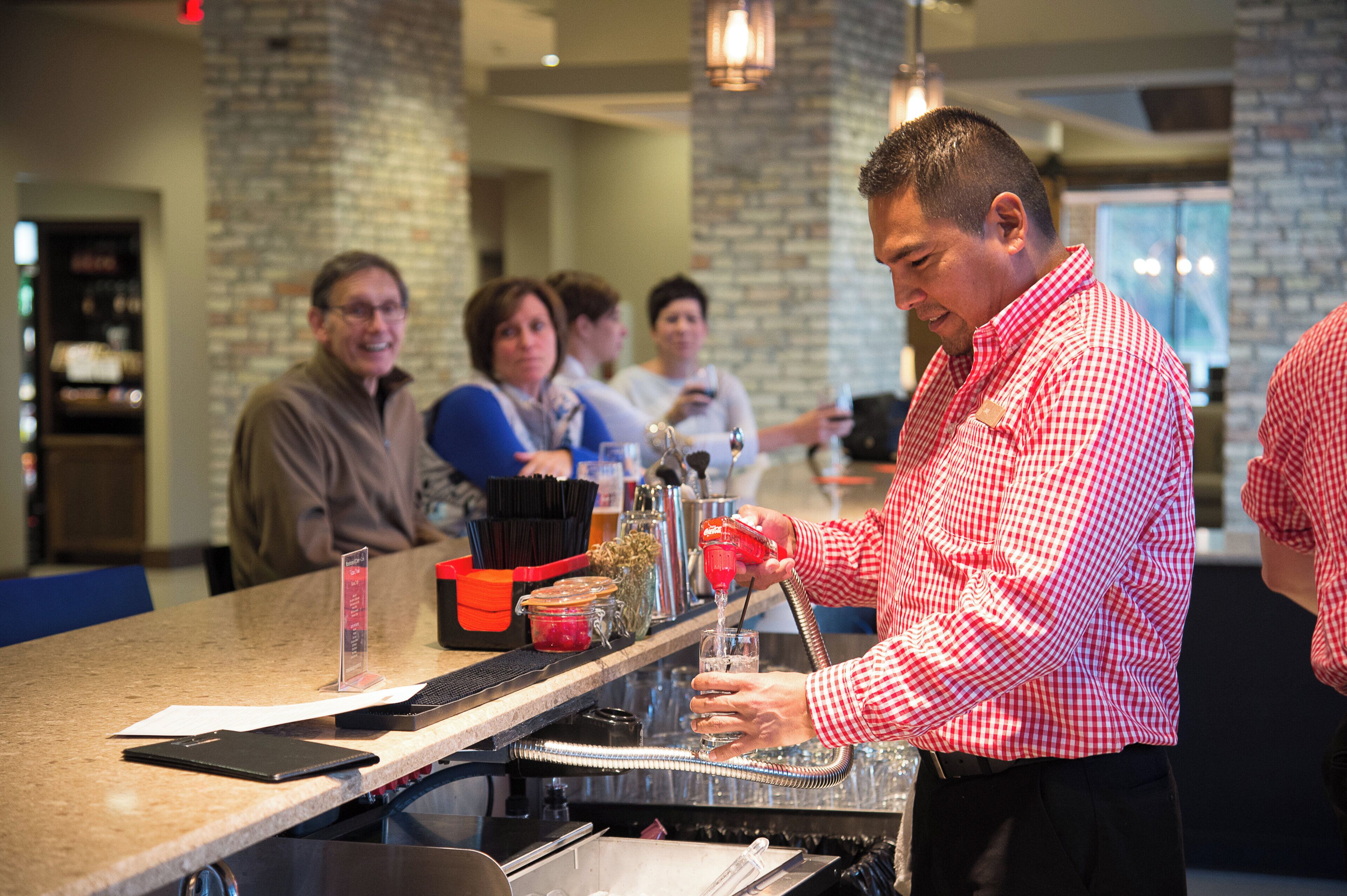 A bar area with a bartender preparing a drink, a menu stand, and a group of people in the background.