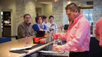 A bar area with a bartender preparing a drink, a menu stand, and a group of people in the background.