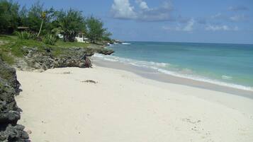 A beach with clear turquoise water, a rocky shoreline, and a few buildings in the background.