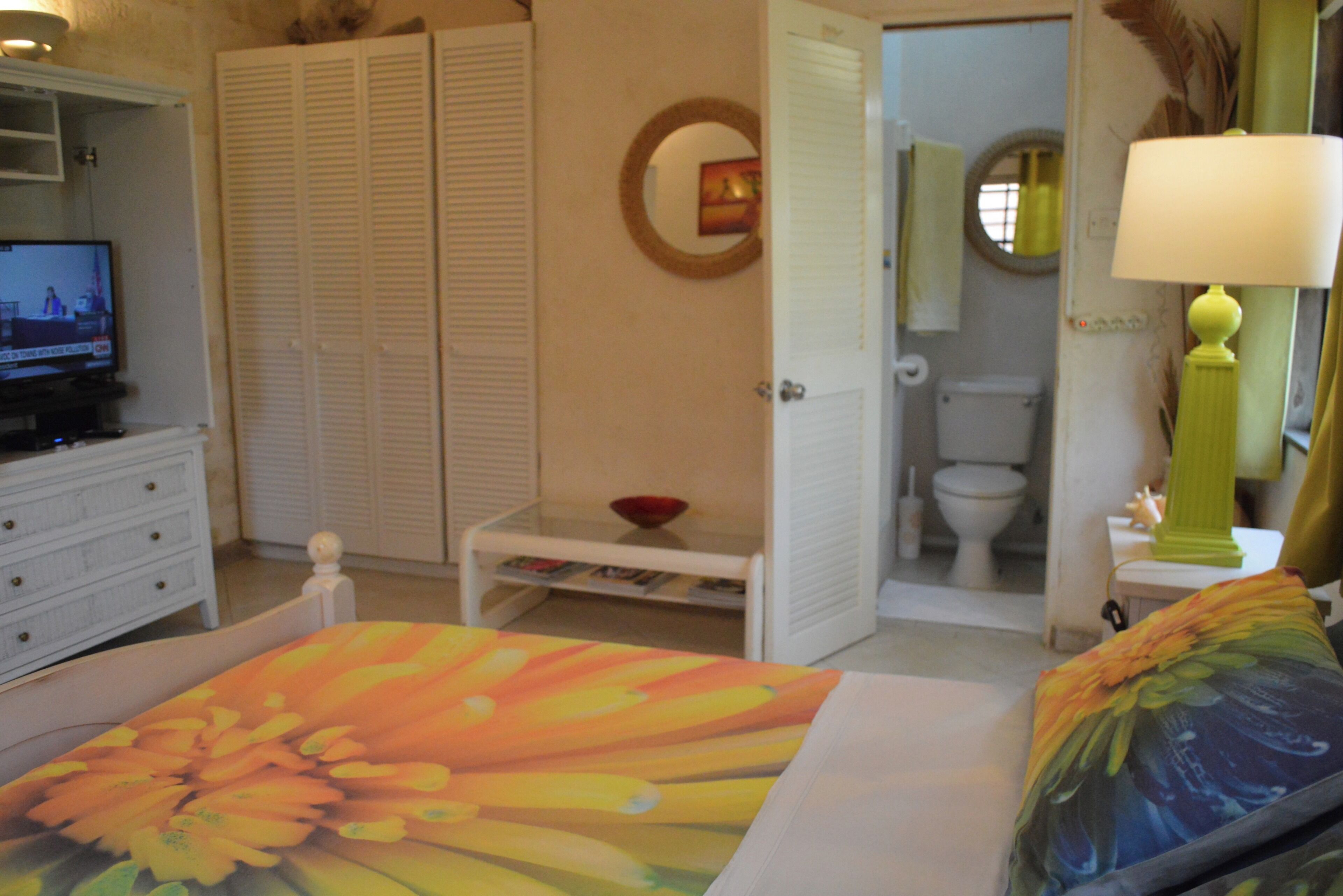 Un dormitorio con una cama, un televisor, un aparador blanco, una mesita con un cuenco, y un baño visible a través de una puerta abierta.
