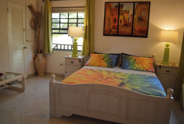 A bedroom with a bed, bedside tables, a window with curtains, a door, and a vase with dried plants. - Yellow Bird Apartment (Silver Sands)