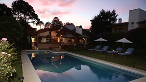 Una piscina con sillas de descanso y sombrillas, rodeada de un césped cuidado y un edificio con iluminación exterior.