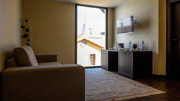 Un salón con sofá, una mesa de centro, un televisor de pantalla plana montado en la pared, un mueble bajo con diversos objetos, y un ventanal que ofrece vistas al exterior.