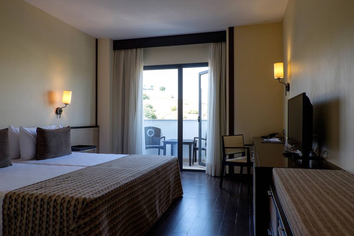 Une chambre d’hôtel avec un grand lit, un bureau avec une chaise, une télévision et un balcon avec des rideaux.
