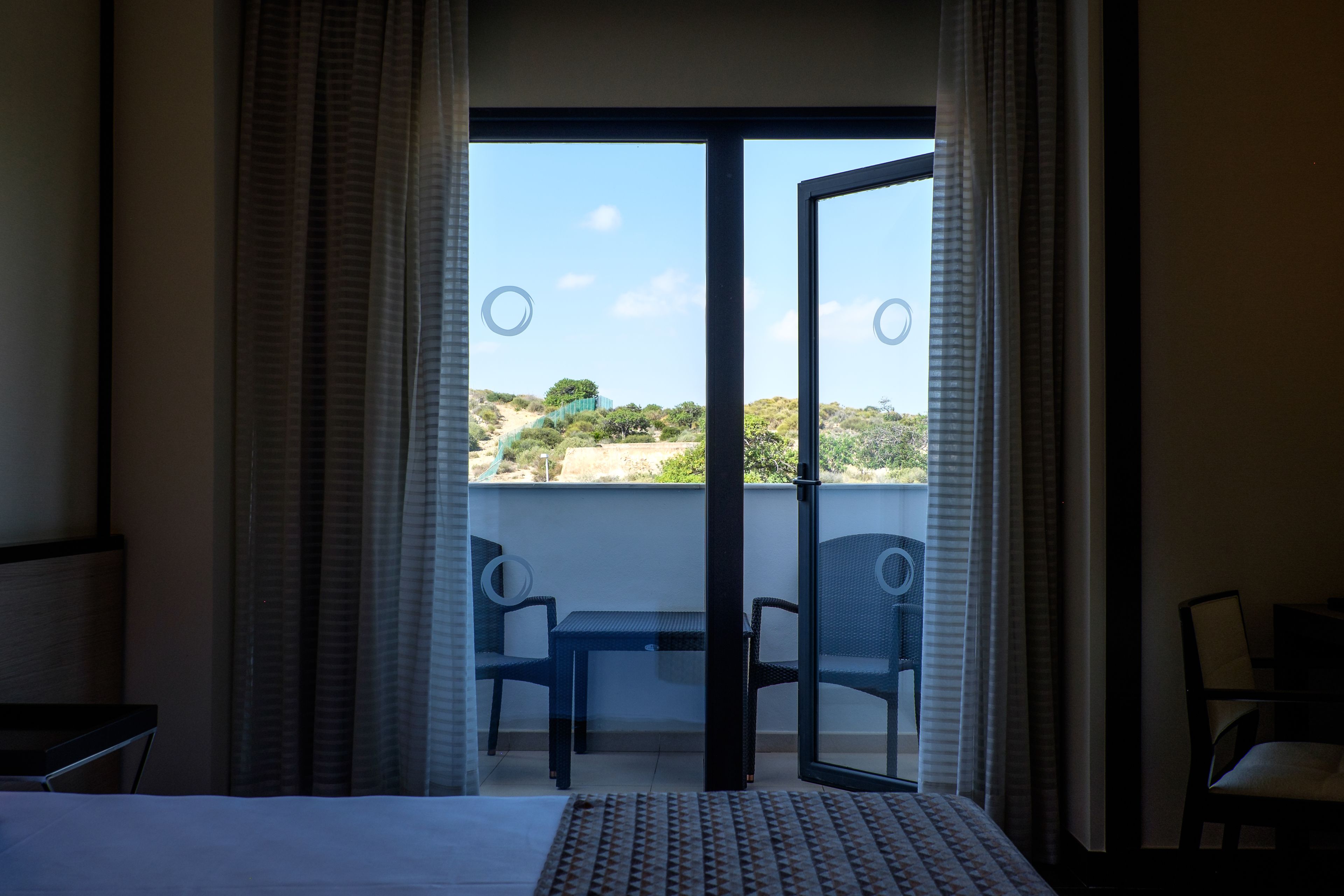 Habitación de hotel con cama, una mesita, dos sillas y un balcón con vistas al paisaje.