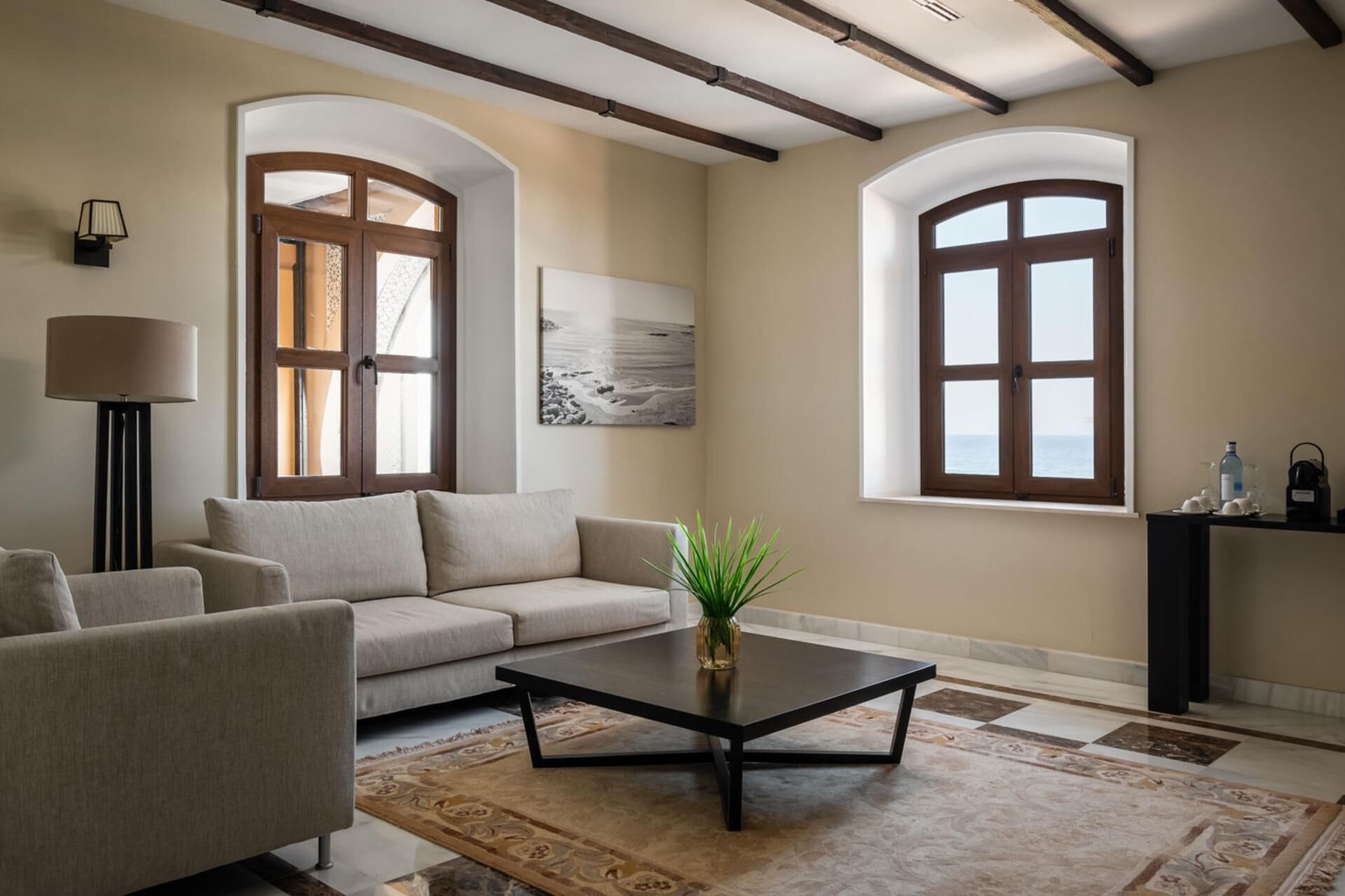Una sala de estar con un sofá, sillones, una mesa de centro y una alfombra. Hay dos ventanas con arcos y un cuadro en la pared.