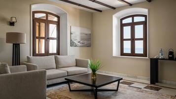 Una sala de estar con un sofá, sillones, una mesa de centro y una alfombra. Hay dos ventanas con arcos y un cuadro en la pared.