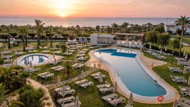A resort with two swimming pools, sun loungers, and a view of the sunset over the sea.