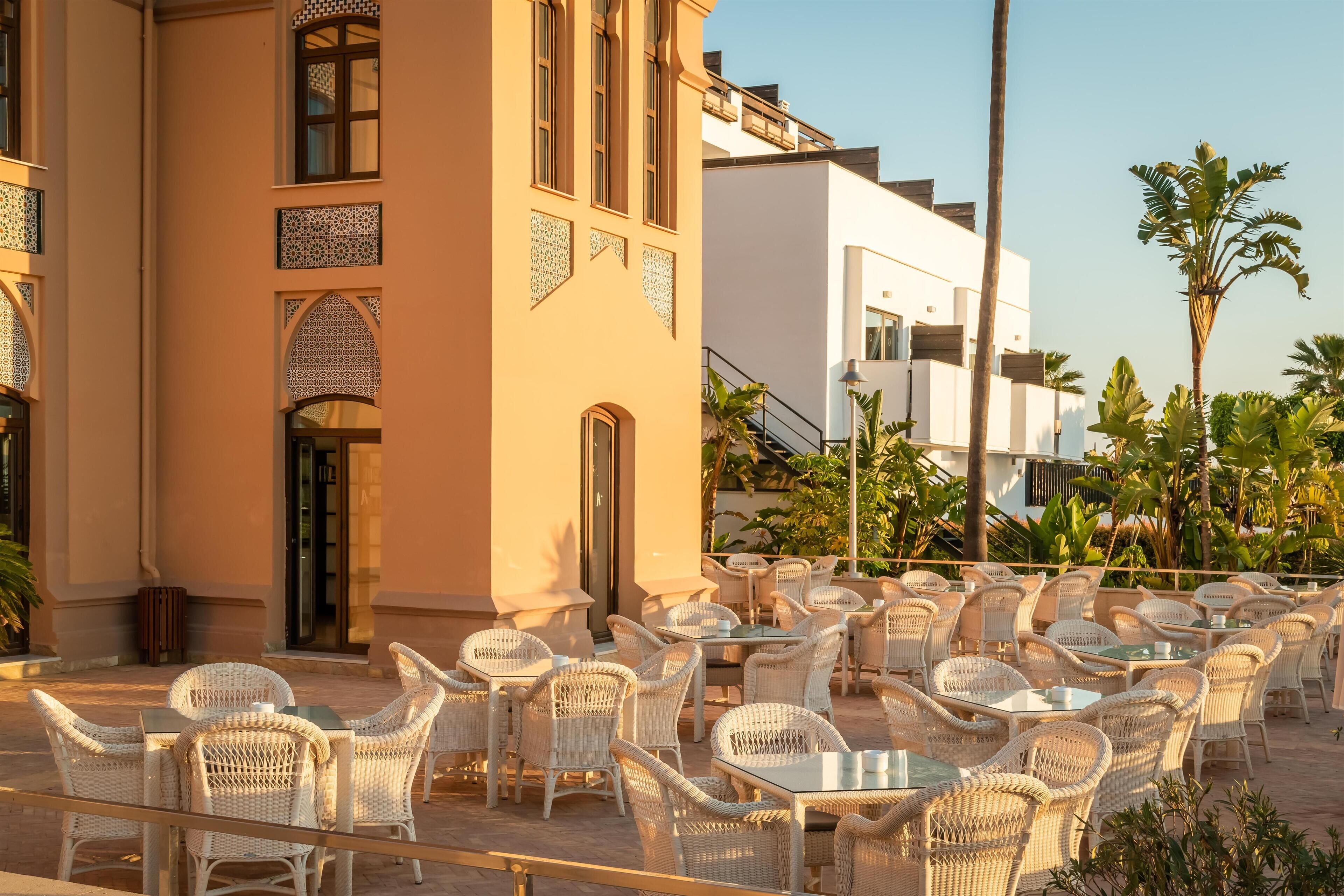 An outdoor seating area with wicker chairs and tables, surrounded by buildings with decorative elements and palm trees.