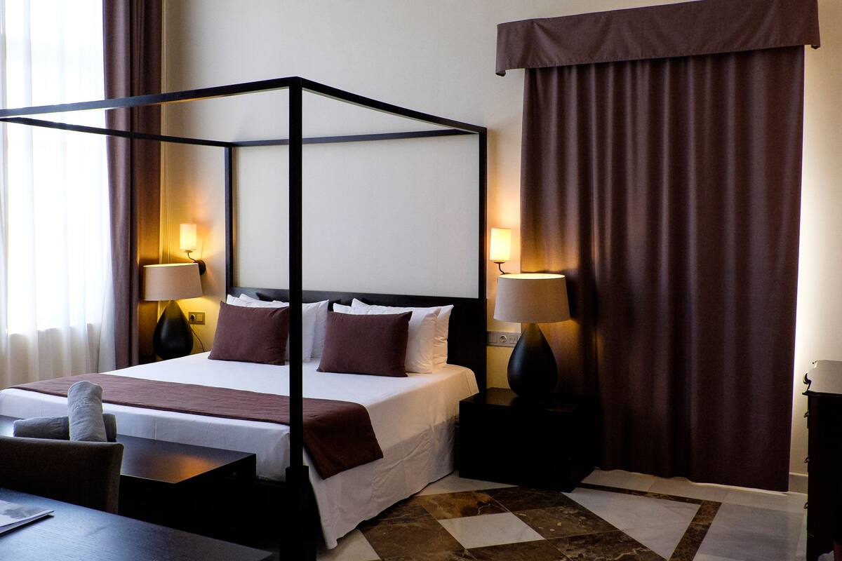 Une chambre d’hôtel avec un lit à baldaquin, deux lampes de chevet, un bureau avec une chaise et un tapis à motifs.