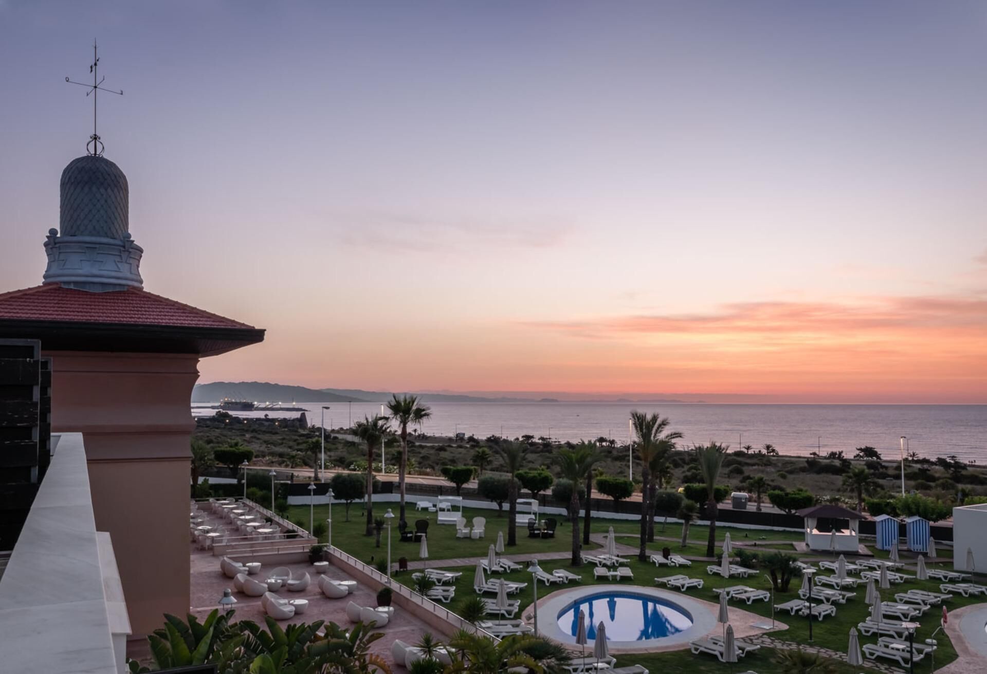 A resort with a pool, lounge chairs, and a view of the ocean at sunset.
