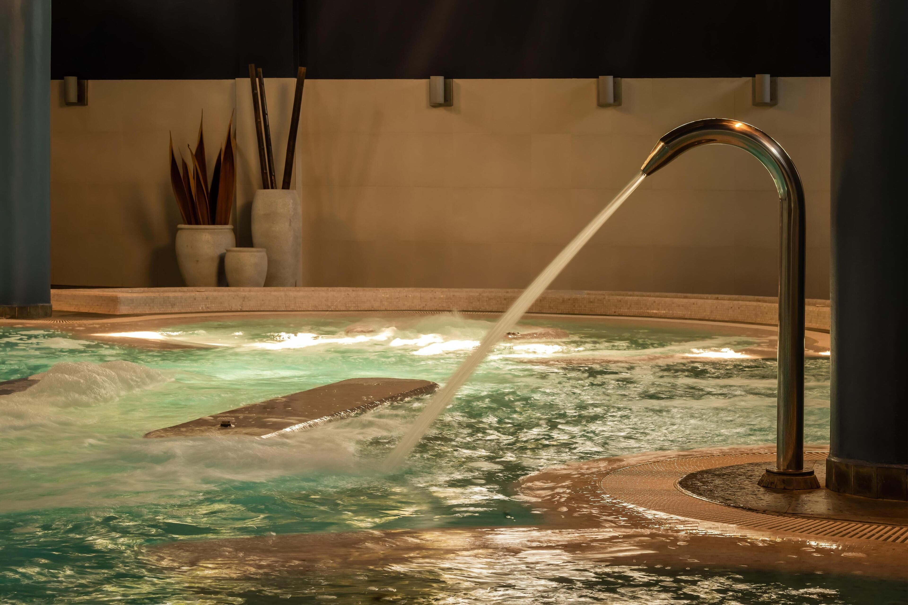 A hot tub with a water spout, surrounded by a tiled floor and a wall with decorative plants.