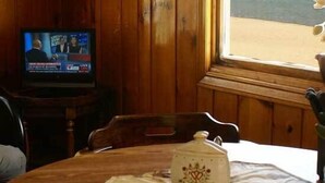 Un interior de madera con un televisor mostrando un noticiero, una mesa con una jarra decorativa y vistas a la playa a través de una ventana.