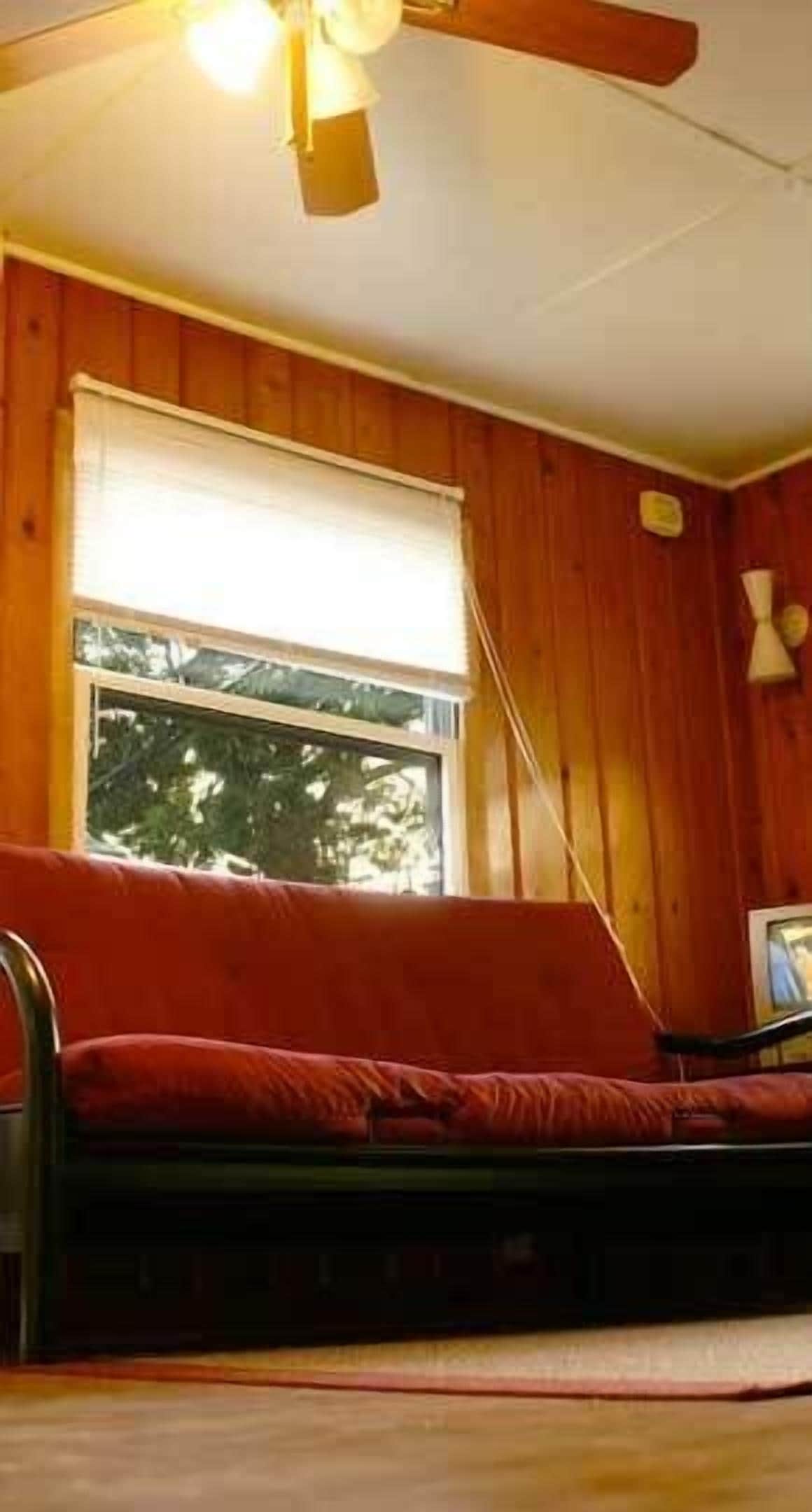 Una habitación con paredes de madera, una ventana con persiana blanca, un sofá rojo y un ventilador de techo.