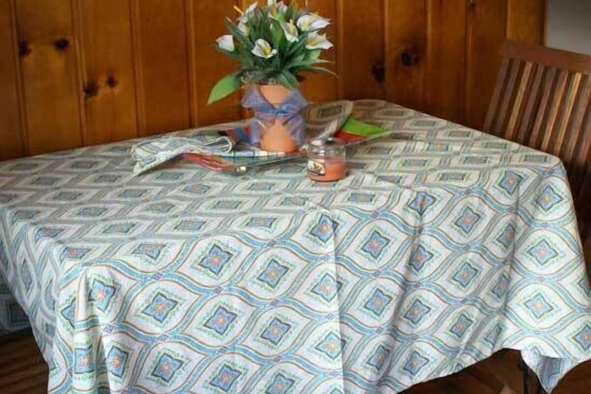 A wooden table with a patterned tablecloth, a vase with flowers, a glass with a drink, and a chair.