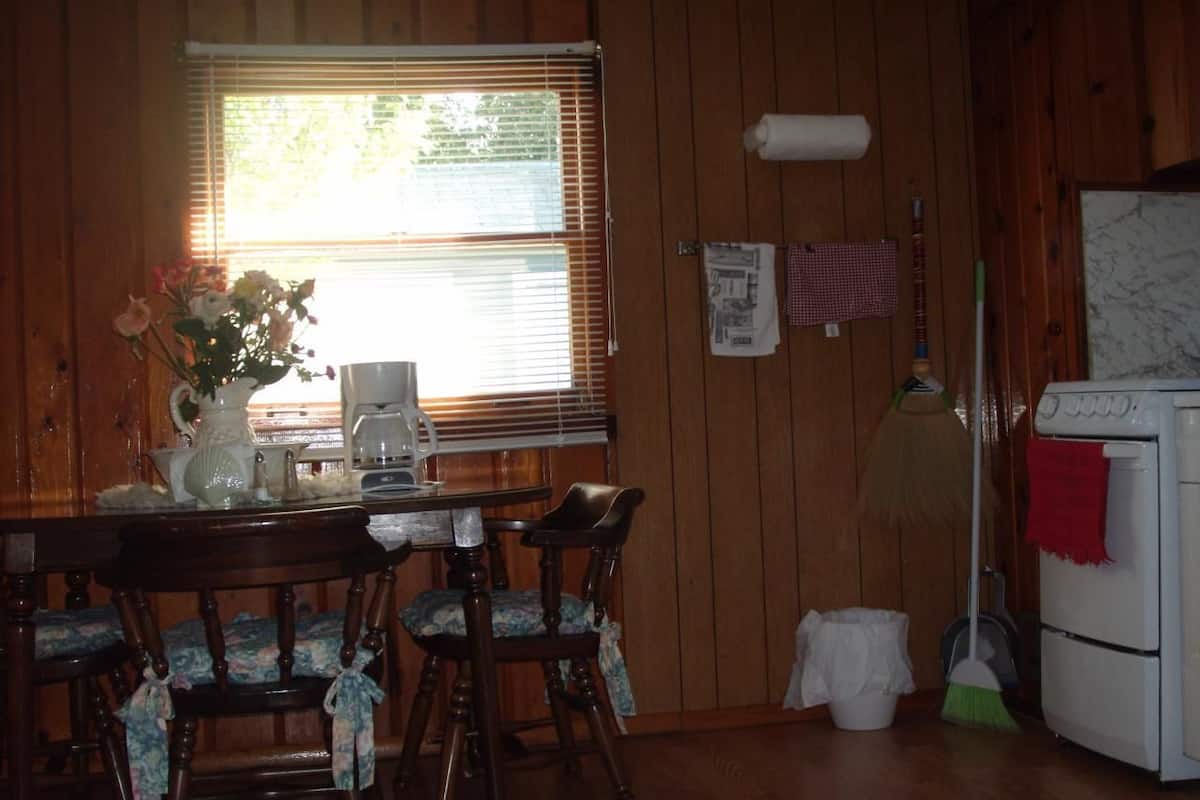 A wooden-paneled room with a dining table, chairs, a coffee maker, a broom, and a stove.