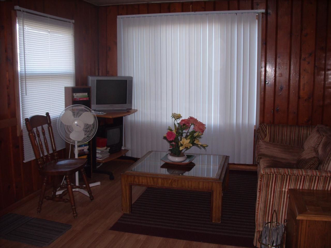 Una sala de estar con televisión, un ventilador, una mesa de centro con flores, un sofá a rayas y una silla de madera.