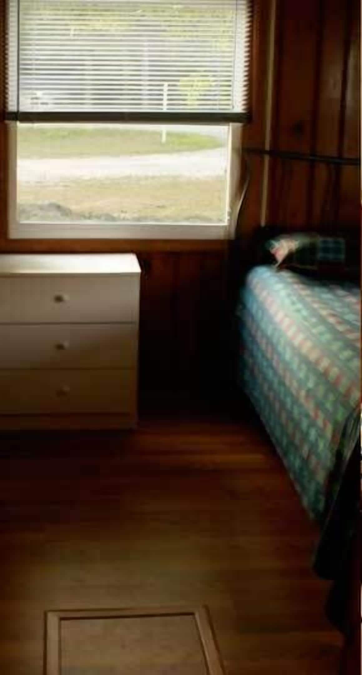 A room with a wooden floor, a bed with a patterned bedspread, a dresser with drawers, and a window with blinds.
