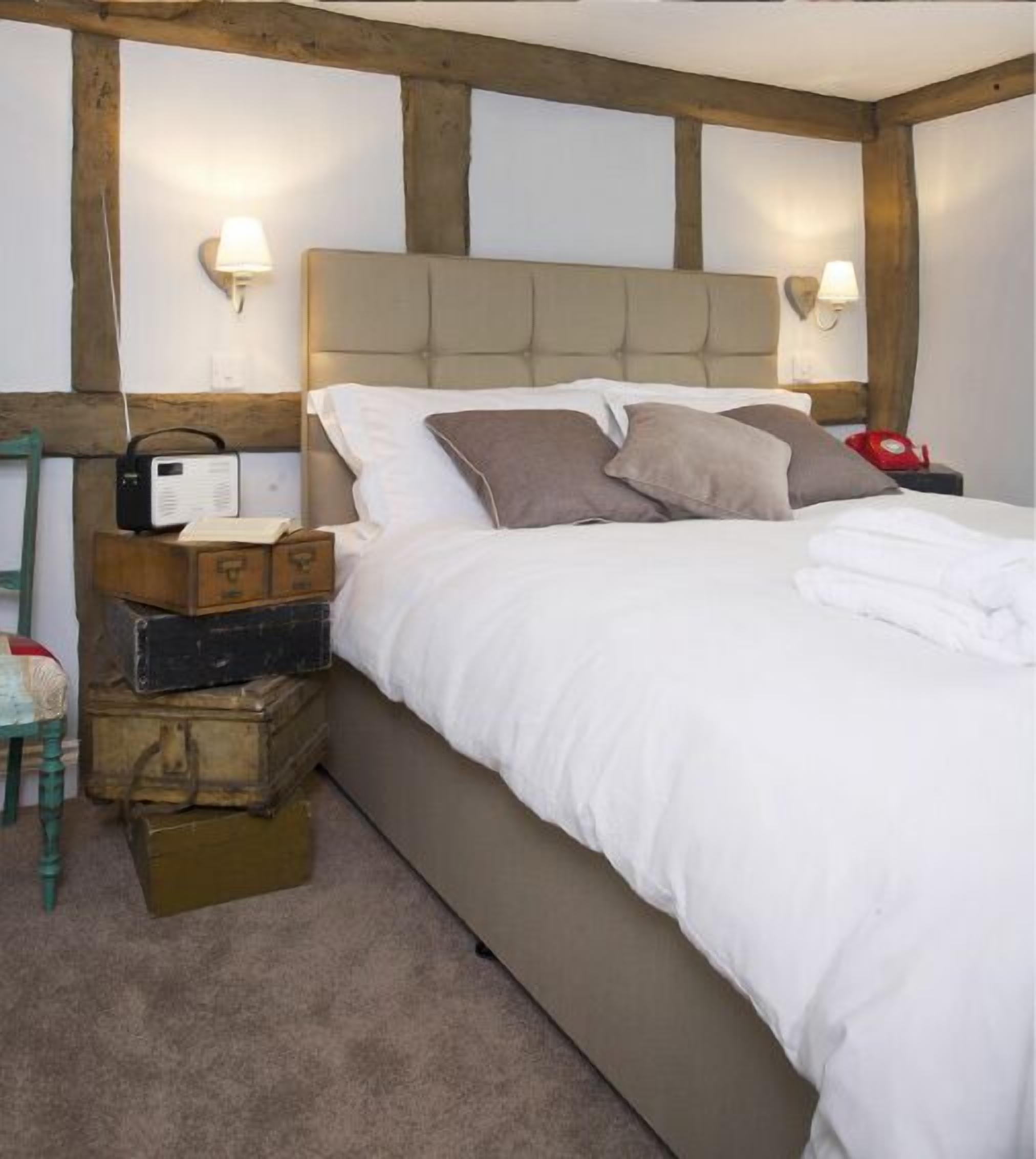 Une chambre à coucher avec un lit, une tête de lit en bois, deux lampes de chevet, une radio et des malles empilées.