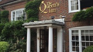 A brick building with a sign reading "The Queen's Inn." There is a covered outdoor seating area with tables and chairs. - The Queen's Inn (Cranbrook)