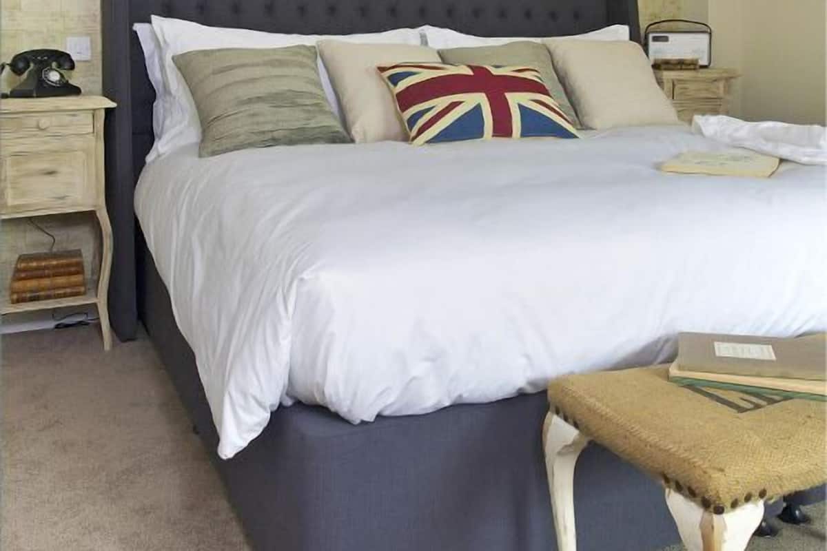 Un dormitorio con cabecera oscura, ropa de cama blanca y una almohada con la bandera Union Jack. Hay dos lámparas de pared con cuernos de ciervo, una mesita de noche con teléfono giratorio y un venado enmarcados en la pared.