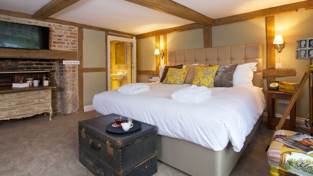 A bedroom with a large bed, a wooden dresser, a television mounted on a brick wall, and a suitcase on a table. - The Queen's Inn (Cranbrook)