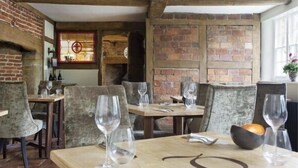 A rustic dining area with brick walls, wooden beams, and a stone fireplace. Tables are set with wine glasses and a bread basket. - The Queen's Inn (Cranbrook)