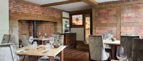 A rustic dining area with brick walls, wooden beams, and a fireplace. Tables are set with wine glasses and cutlery.