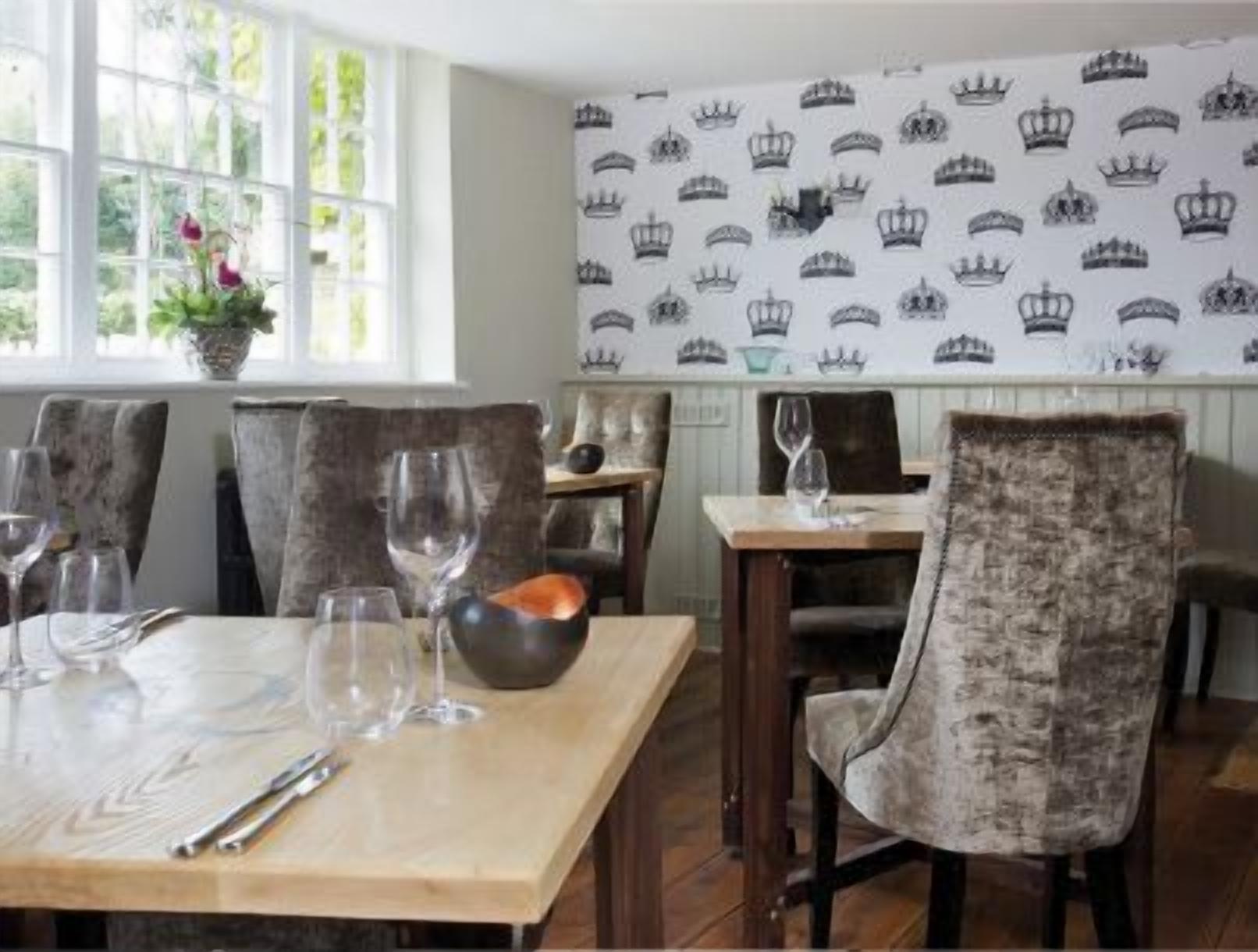 A dining area with a table set for two, featuring a patterned wallpaper with crown designs, upholstered chairs, and a window with a view of greenery.