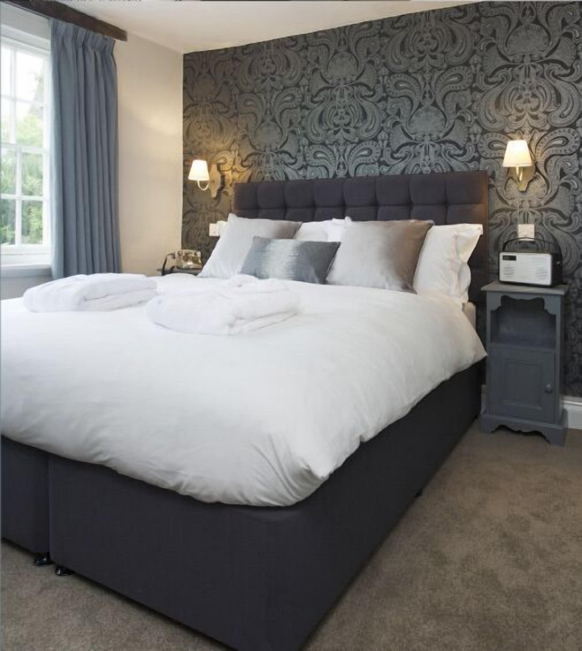 A neatly made bed with white linens and multiple pillows, a dark headboard, and a wall with a patterned wallpaper.