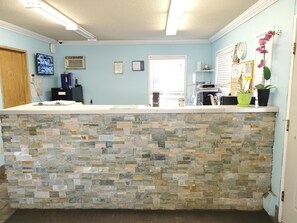 A reception area with a stone counter, a wooden door, a security camera, a television, and a window with blinds. - Stars Inn & Suites Building B (Fort Saskatchewan)