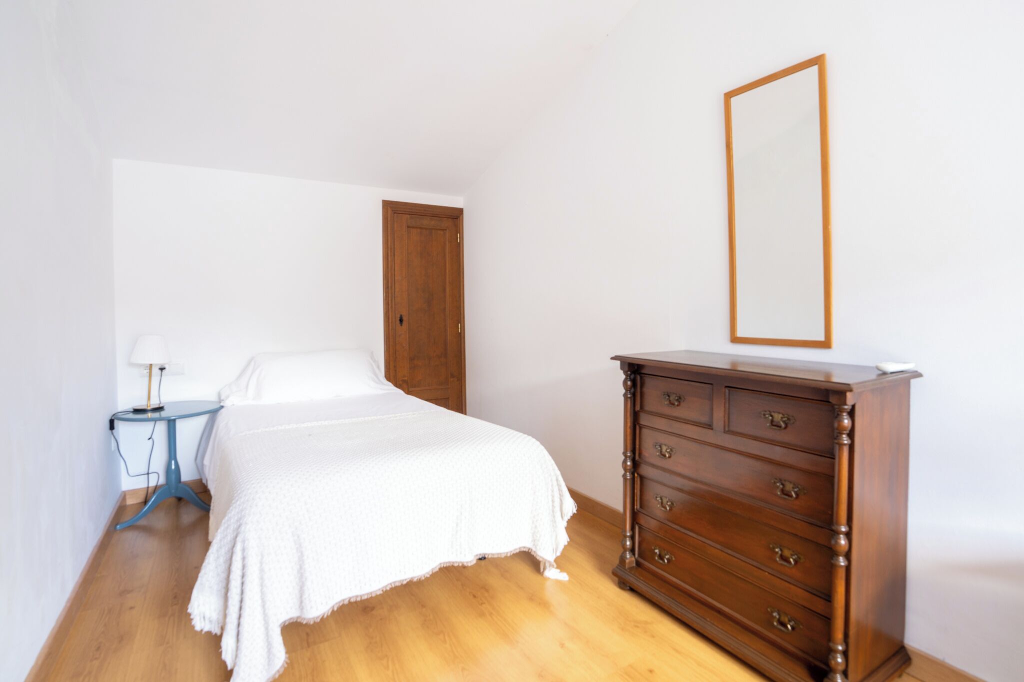 A bedroom with a bed, a dresser, a nightstand, a wooden door, and a mirror on the wall.