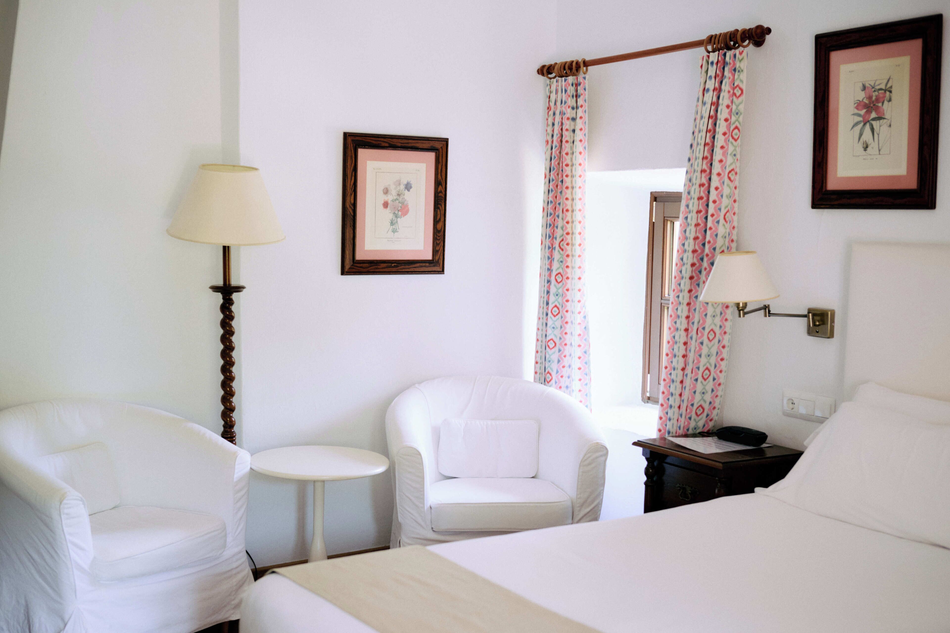 Quarto de hotel com uma cama, duas cadeiras brancas, uma mesinha redonda, um criado-mudo com abajur e uma janela com cortinas estampadas.