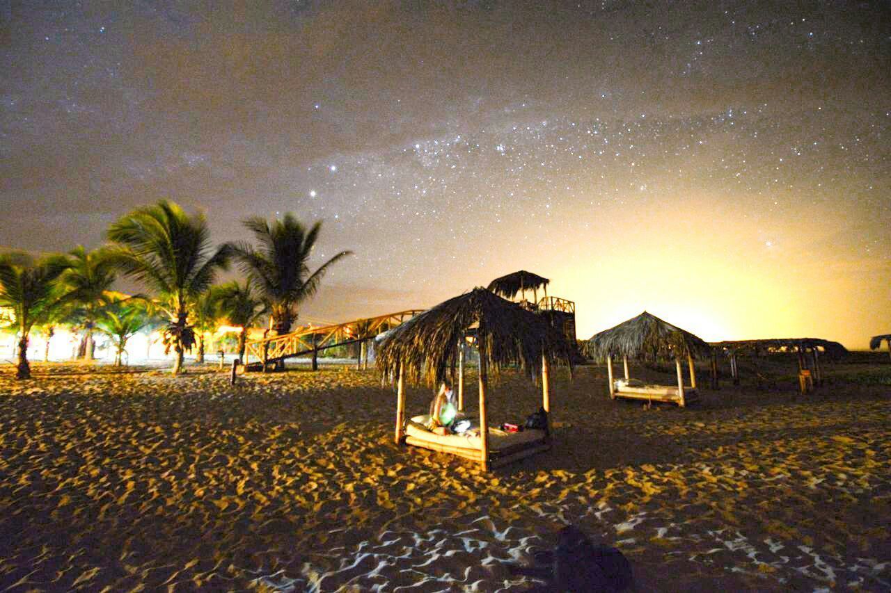 Une plage bordée de huttes aux toits de chaume et de palmiers, sous un ciel étoilé.