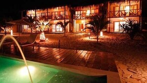 Vista nocturna de un resort con piscina, sillas de descanso y un edificio de varios niveles.