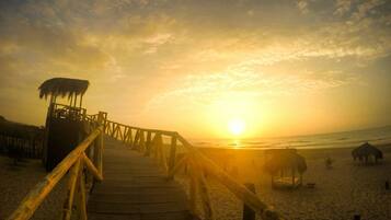 Un paseo marítimo de madera que lleva a una playa con construcciones de techo de paja.