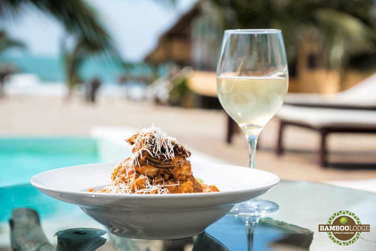 Una mesa de comedor con un plato de pasta y una copa de vino blanco, con el mar y la playa al fondo.