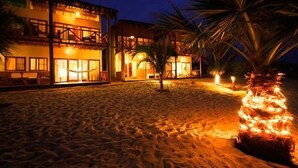 Un hotel frente a la playa con varios balcones, un patio de arena y palmeras.