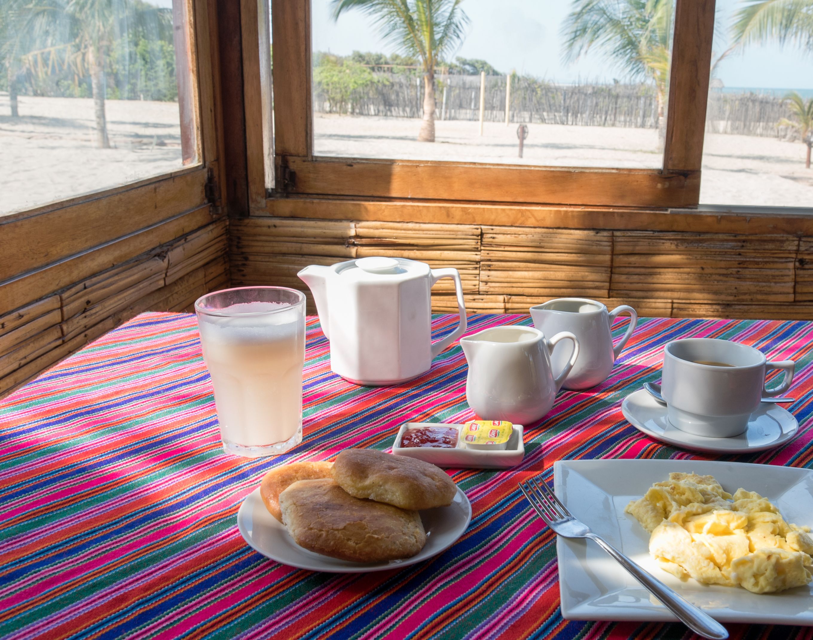 A breakfast table with a striped tablecloth, a pitcher, cups, a plate of pastries, and a glass of beverage.