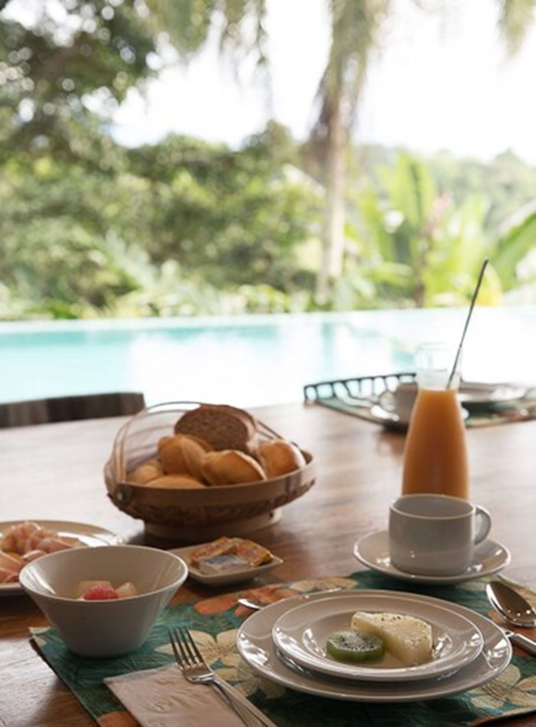A breakfast setting with a pool view, including a bowl of fruit, a plate with a dessert, a cup of coffee, and a glass of orange juice.