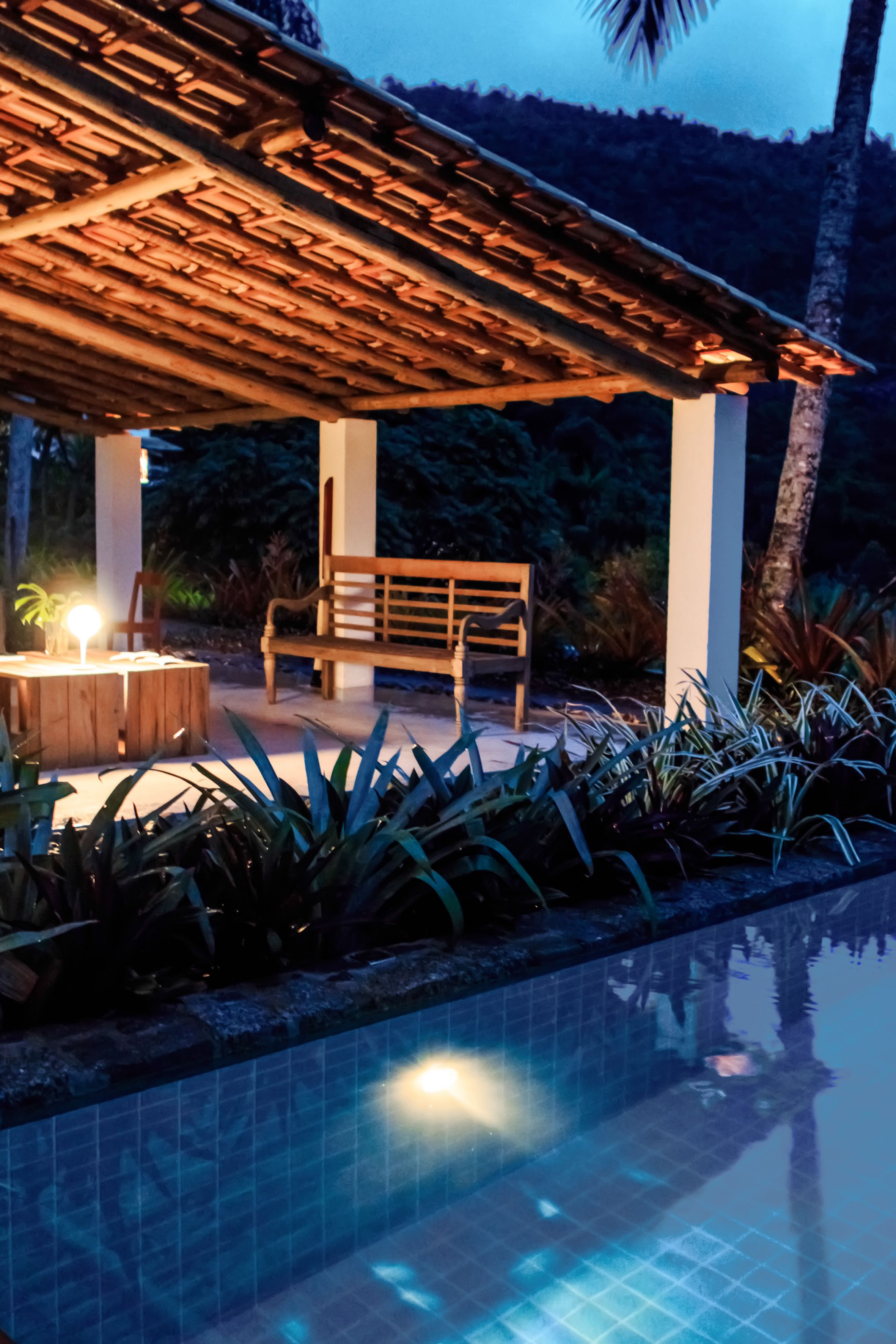 Zona de piscina con banco de madera y mesa bajo una pérgola, rodeada de plantas tropicales.