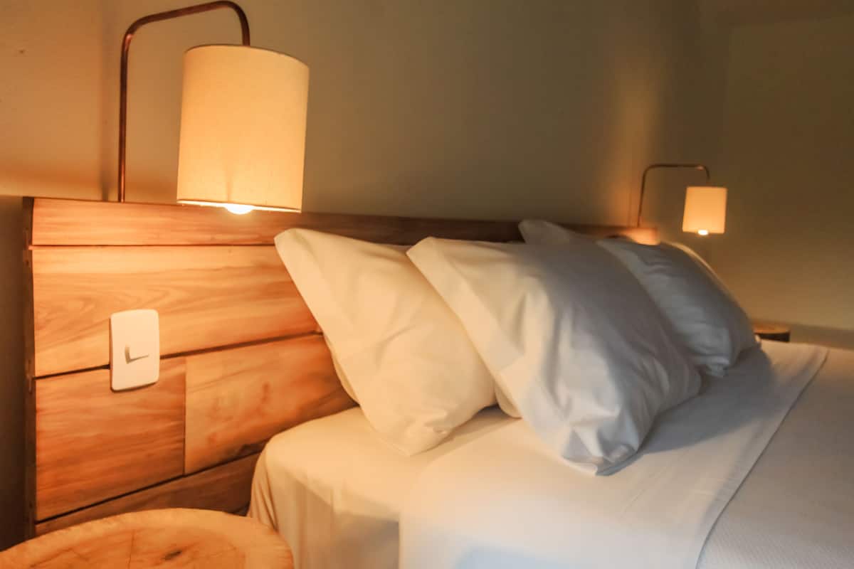 Una cama bien hecha con sábanas y almohadillas blancas, un cabecero de madera con un reloj, y dos lámparas de pared que brindan una luz suave.