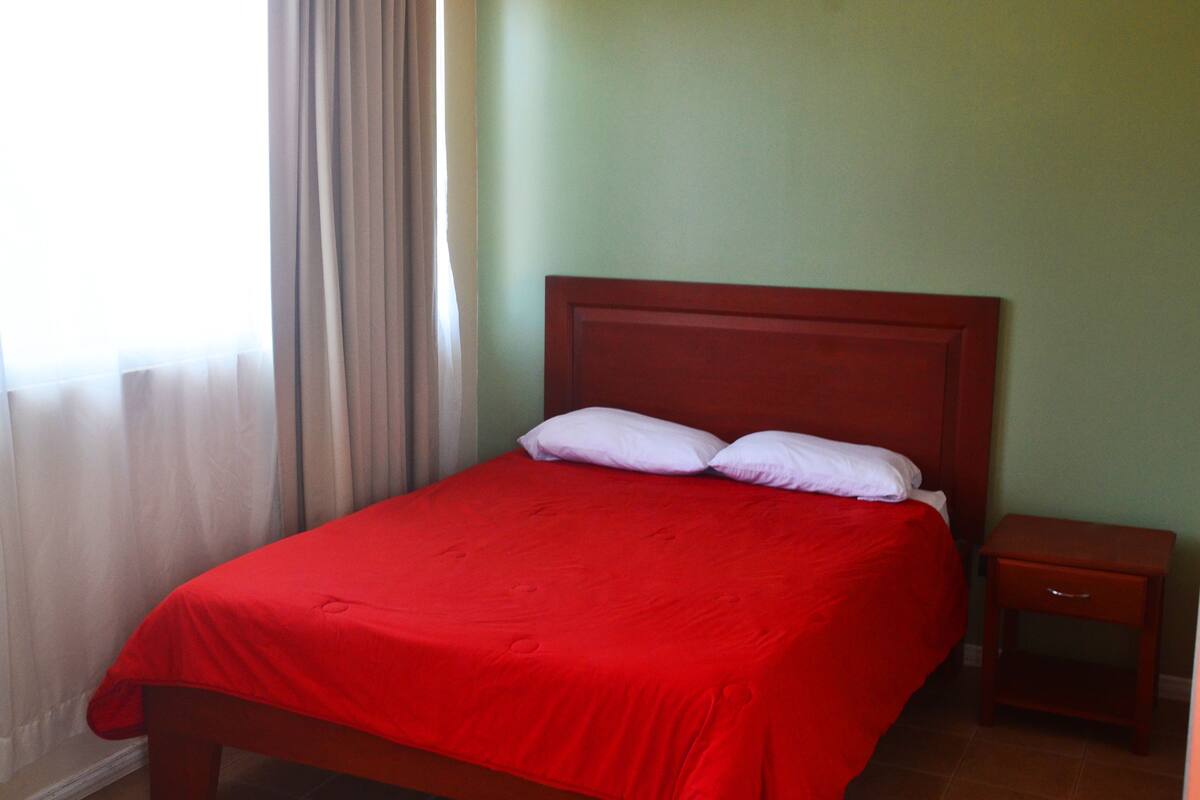 A neatly made bed with a red bedspread and white pillows in a room with green walls and a wooden nightstand.