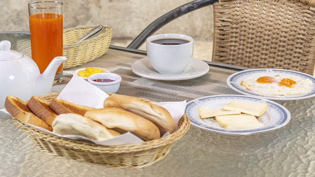Un desayuno con una silla de mimbre, un vaso de jugo de naranja, una tetera, una taza, platos con huevos, tostadas y una cesta de pan.